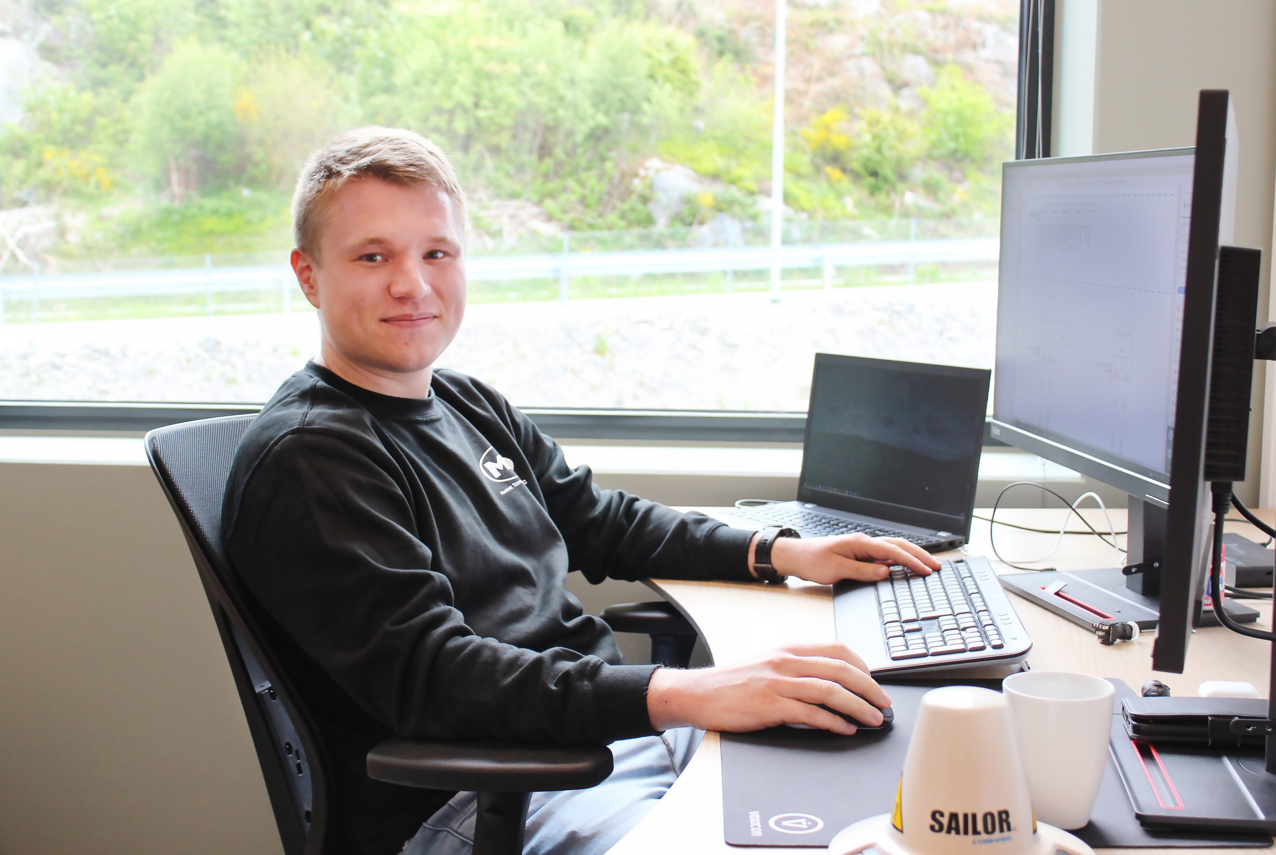 JOn SIvert Andreassen på kontoret sitt iført sort genser med MT-logo. Han sitter foran pcen sin, og i bakgrunnen kan man se grønne busker gjennom vinduet.
