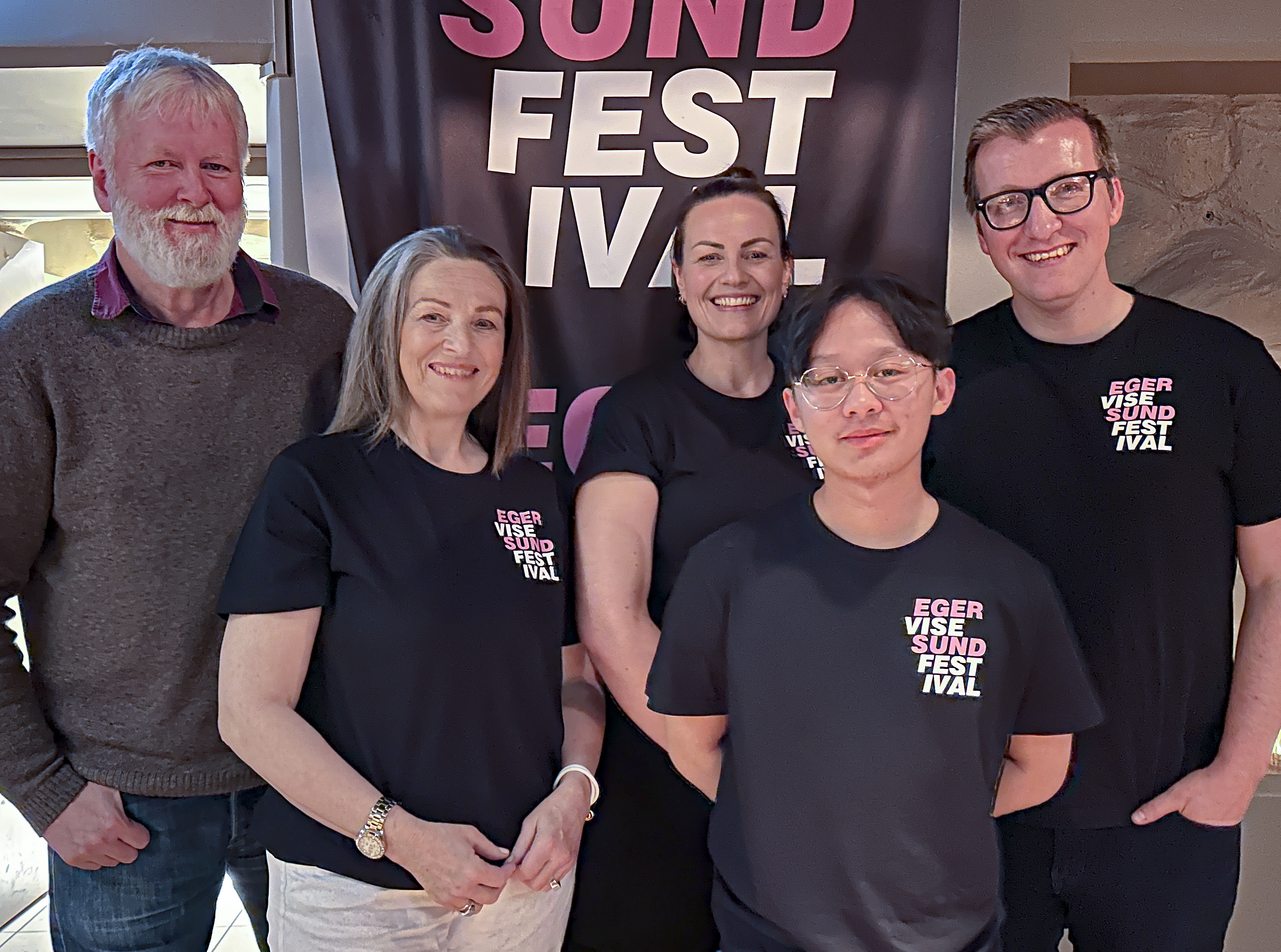 Fem medlemmer av Egersund Visefestival-komité. Flesteparten er iført mørke t-skjorter som viser den nye logoen laget til festivalen. Logoen er lys rosa og hvit.