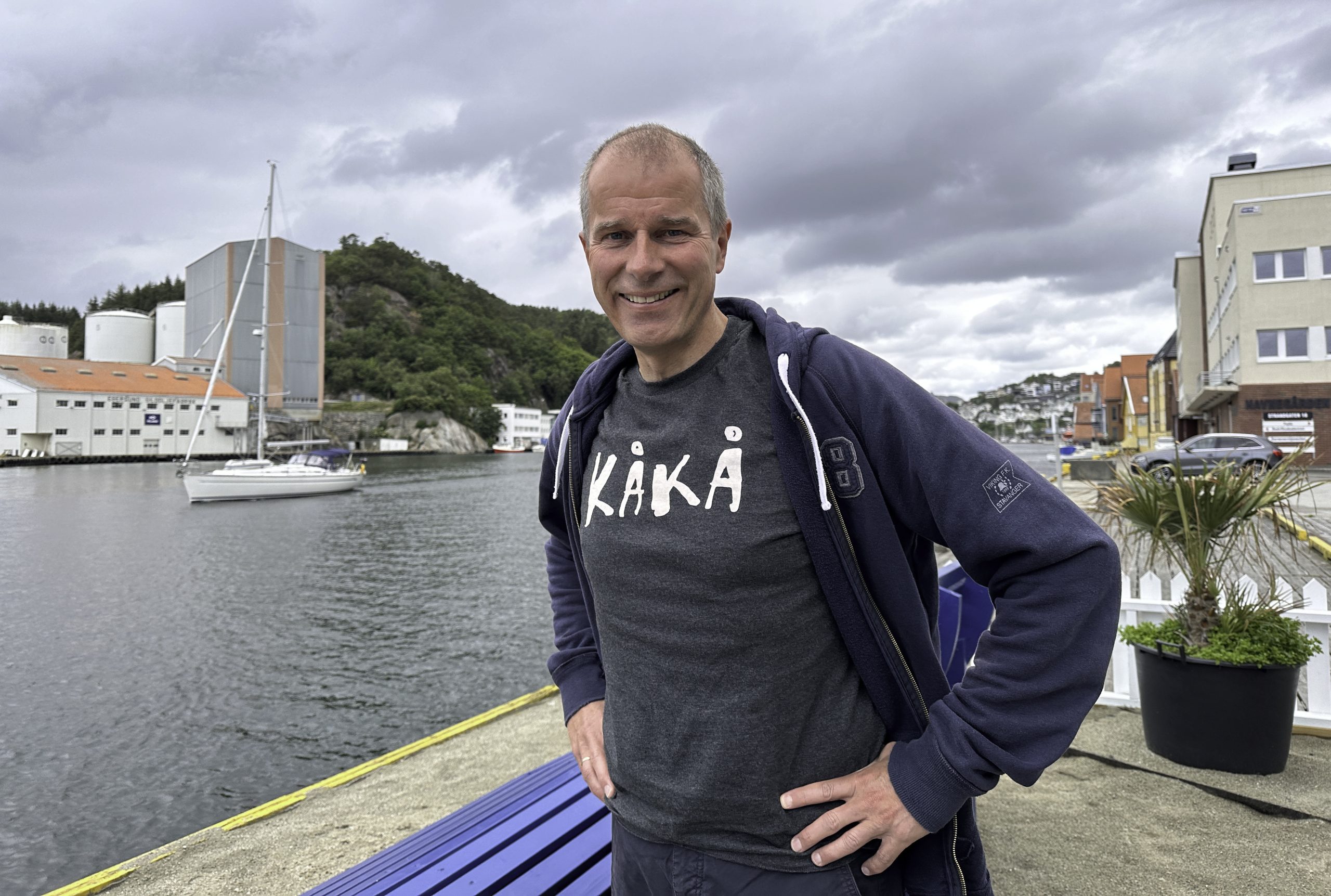 Cornelius Middelthon står på Bredbenken strand i Egersund. Han har på seg en blågrå t-skjorte med teksten KÅKÅ. I bakgrunnen kjører en seilbåt.