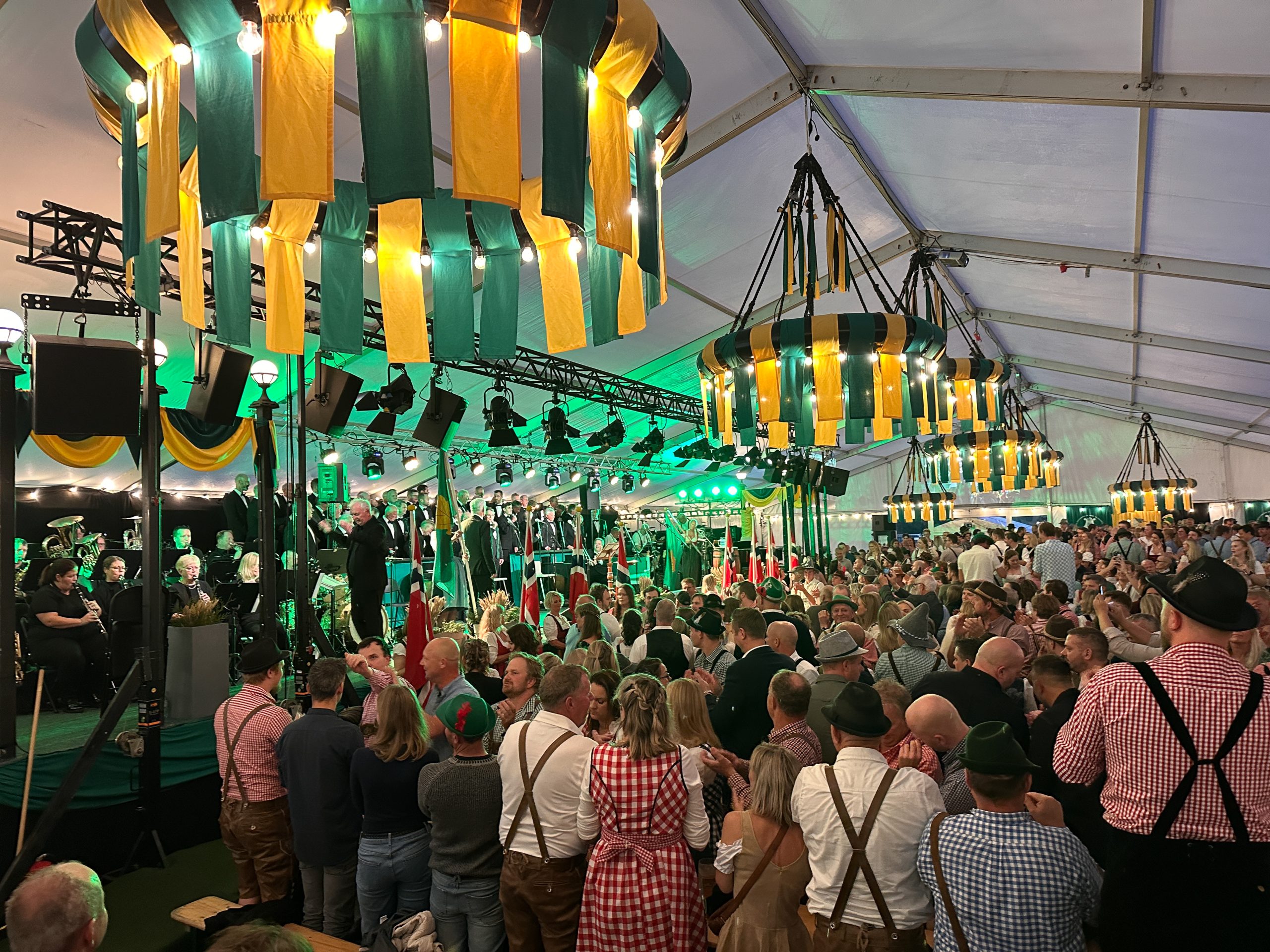 innsiden av oktoberfestteltet. Vi ser de tradisjonelle lampeskjermene i grønt og gult og mange mennesker ikledd dirndl og lederhosen