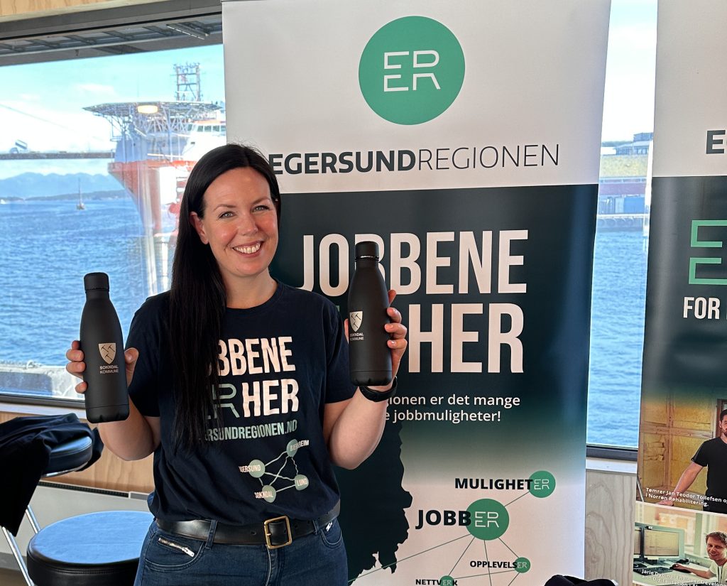 Helene Holm avbildet i t-skjorte med påskriften "jobbene er her", Egersundregionen-logo og kommunene Eigersund, SOkndal, Bjerkreim og SOkndal. Hun holder to svarte flasker i hendene med logo til Sokndal kommune.